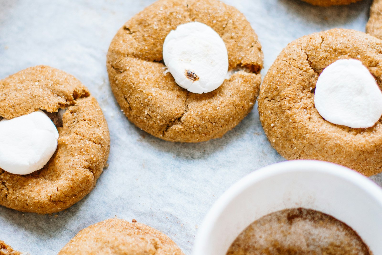 Chocolate thumbprint cookies being filled with white chocolate peppermint ganache from a small spoon, with a bowl of crushed candy canes beside the baking sheet