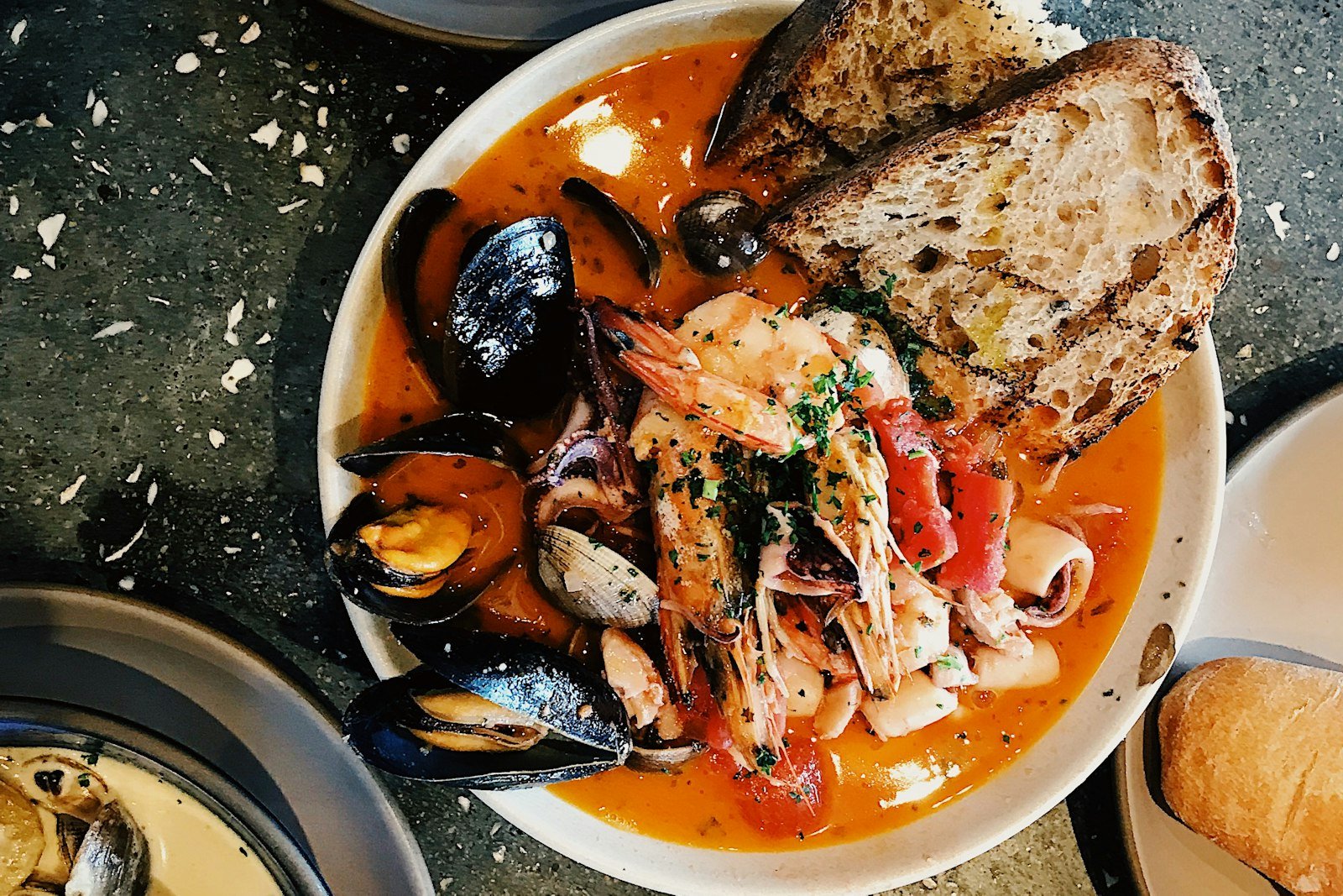 A ladle lifting cioppino from a Dutch oven, showing opened mussels, a pink shrimp and a chunk of flaky white fish in tomato broth