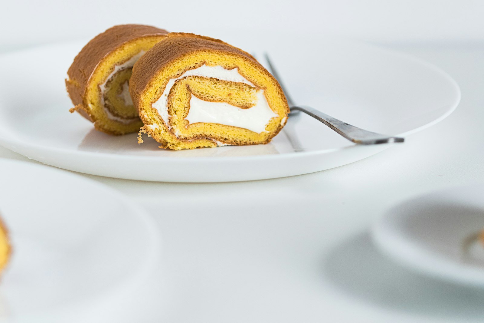 A pumpkin sponge cake partly unrolled on a powdered-sugar-dusted kitchen towel, with cream cheese filling being spread across its surface with an offset spatula