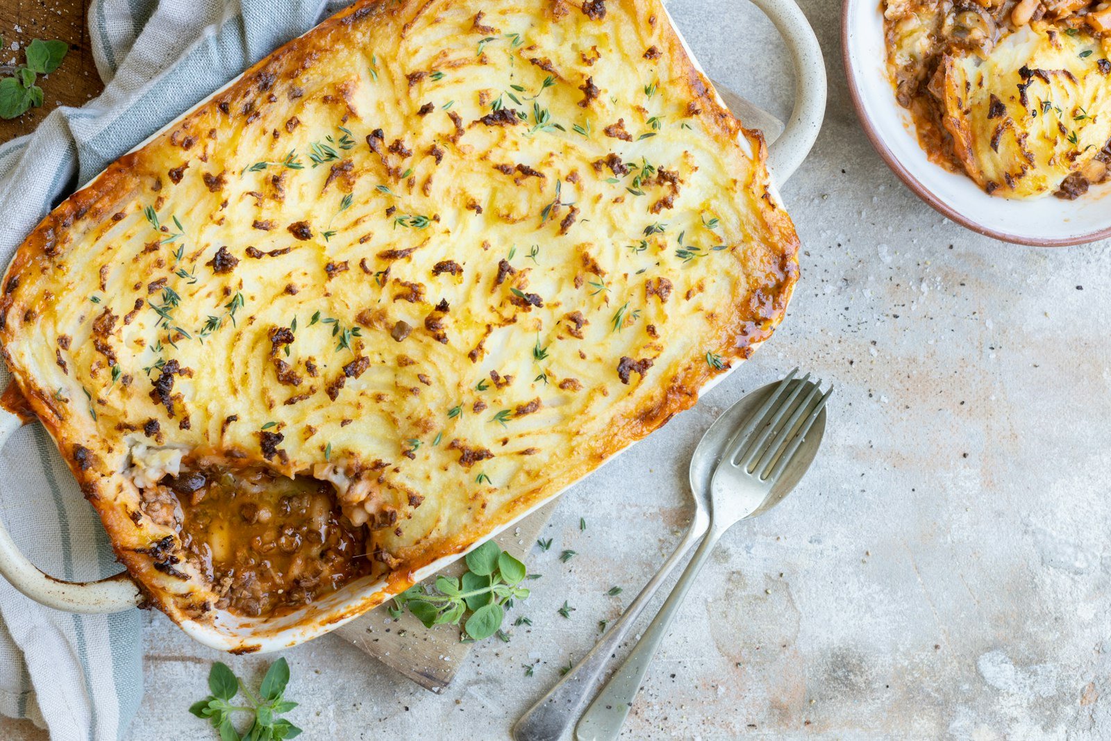 A serving spoon lifting a portion of shepherds pie from the baking dish, showing the golden mashed potato crust and the saucy ground lamb, carrot and pea filling underneath